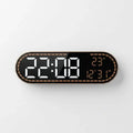 A modern digital wall clock with an elongated rounded rectangular shape, featuring a black face, a metallic bronze border with a dotted pattern, and white LED time display alongside orange LED temperature and date display.