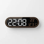 A modern digital wall clock with an elongated rounded rectangular shape, featuring a black face, a metallic bronze border with a dotted pattern, and white LED time display alongside orange LED temperature and date display.