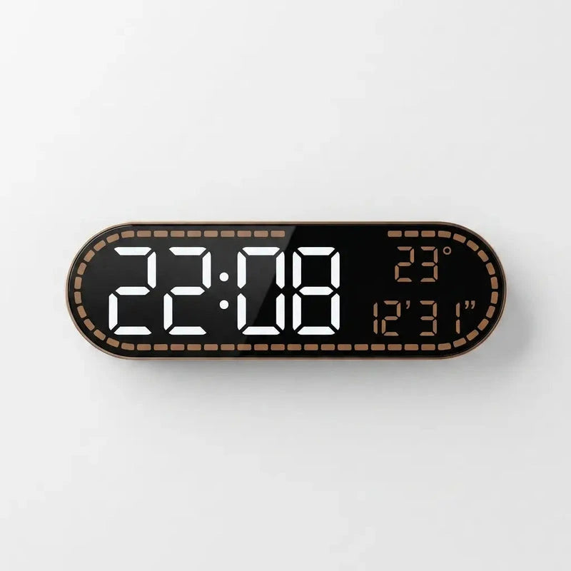 A modern digital wall clock with an elongated rounded rectangular shape, featuring a black face, a metallic bronze border with a dotted pattern, and white LED time display alongside orange LED temperature and date display.