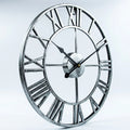A large round wall clock with an open Roman numeral design, featuring a distressed silver metal frame and black hands, shown against a light gray studio background.