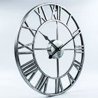 A large round wall clock with an open Roman numeral design, featuring a distressed silver metal frame and black hands, shown against a light gray studio background.